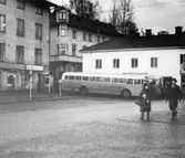 Ett gående par passerar vid Gamla Torget i Kvarnbyn, Mölndal, på 1960-talet. I bakgrunden till vänster ses en telefonkiosk utanför tvätteriet ALBA-tvätt i huset Kvarnbygatan 43. I bakgrunden i mitten ses Hedströms hus med beteckningen Roten F8 och F9. I bakgrunden till höger ses två bussar utanför huset Kvarnbygatan 45.