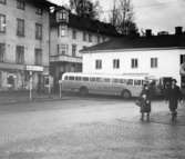 Ett gående par passerar vid Gamla Torget i Kvarnbyn, Mölndal, på 1960-talet. I bakgrunden till vänster ses en telefonkiosk utanför tvätteriet ALBA-tvätt i huset Kvarnbygatan 43. I bakgrunden i mitten ses Hedströms hus med beteckningen Roten F8 och F9. I bakgrunden till höger ses två bussar utanför huset Kvarnbygatan 45.