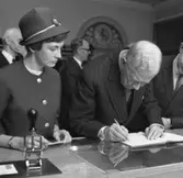 Jubileumshedersgäst var dåvarande kung Gustaf VI Adolf. Här skriver
kungen i gästboken och sätter sin namnteckning på ett museibrevkort,
under överinseende av postvärdinnan Inger Svärd.