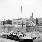 En liten båt vid kajen gör reklam för Radio Nord. Stationen sände från ett fartyg, M/S Bon Jour, förankrat på internationellt vatten utanför Stockholms skärgård med premiären den 8 mars 1961 och stationen upphörde den 30 juni 1962.