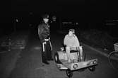 Kvarterspolis Ola Candefors, Batong-Ola kallad, ses tillsammans med en pojke som sitter på något slags fordon utanför Almåsgården i Lindome, år 1983.

För mer information om bilden se under tilläggsinformation.