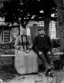 Ett äldre par sittande på en bänk.
Bostadshus i bakgrunden.