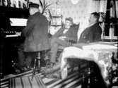 Rumsinteriör, tre män, en av dem spelar piano.