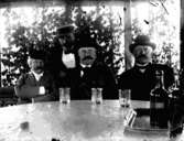 Fyra herrar vid bordet.
Tre dricksglas och bricka med två flaskor på bordet.