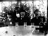 Fyra herrar vid bordet.
Tre dricksglas och bricka med två flaskor på bordet.