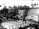 Sågverk, lastning av trävirke i tågvagnar, sex arbetare.
I bakgrunden en banvaktstuga av ålderdomlig modell.