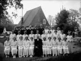 Konfirmander, 23 flickor, 6 pojkar och kyrkoherde Ivar Thorell.
Kvistbro kyrka i bakgrunden.