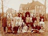 Almby Folkskolan, en och en halvvånings skolbyggnad i bakgrunden.32 skolbarn med lärarinnan fröken Andersson.