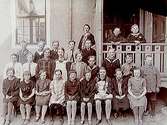 Rynninge folkskola, 25 skolbarn med lärare fröken Mossberg.
Skolbyggnad i bakgrunden.