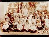 Rynningeskolan, 23 skolbarn med lärarinna fröken Dahlin på skolgården.
Skolbyggnad i bakgrunden.