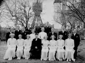18 konfirmander, 9 flickor, 9 pojkar och kyrkoherde Ivar Jäderlund.
Glanshammars kyrka och klockstapel i bakgrunden.