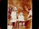 En kvinna med två småbarn, en av dem sitter på en stol.
Th. Rosell