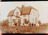 En och en halvvånings villa med frontespis, burspråk, glasad veranda och brutet tak.
10 personer framför villan.
P.G. Broström