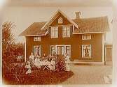 En och en halvvånings bostadshus med takhuv, separat köksingång. Grupp, 7 personer vid kaffebordet framför huset.
Albin Karlsson
