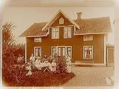 En och en halvvånings bostadshus med takhuv, separat köksingång. Grupp, 7 personer vid kaffebordet framför huset.
Albin Karlsson
