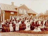 En och en halvvånings bostadshus med takhuv och förstubro i snickarglädje.
Bröllop. Brudpar och bröllopsgäster framför huset.
Aug. Andersson