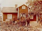 Envånings bostadshus med trapphus.
En kvinna och två män framför huset.
Alfred Vase
