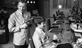 Första spad, Polis i skolorna 13 okt 1967

På en frisörsalong ser man en frisör som håller en hårfläkt
i handen, och en man sitter i en stol. Det finns också fyra stolar till och flera handfat, och hårprodukter.