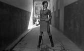 Modenummer Sand 30 april 1968

Mannikäng i dräkt med vit krage och skinnstövlar står i en portingång med grind en som är öppen. Bakom henne ser man Stortorget där bilar står parkerade, och en kvinna med barnvagn.