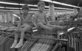 Mannekängande på Domus 21 mars 1968

En flicka och en pojke sitter på en glasskiva på Domus klädavdelning. Flickan har en klänning, och pojken har kortbyxor och en kortärmad jacka på sig.