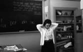 Kopparberg skola, 1 mars 1967.

I ett klassrum står en lärarinna vid en kateder. Hon är klädd i en vit blus, vit kofta och svart kjol.  Hon håller på att ordna sin frisyr.


Fröken Karin Flodström