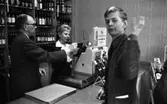 Stoppljus på systemet, 3 juni 1965

En yngre man handlar på Systembolaget. Han betjänas av en kassörska som står bakom disken. Hon håller ynglingens plånbok med identitetskort i handen. Bredvid kassörskan bakom disken står en man och betraktar tillsammans med henne den unge mannens legitimation. Han är klädd i mörk kostym och bär glasögon. I bakgrunden syns hyllor med vin- och ölflaskor. Även vid kassaapparaten står vinflaskor.
Kassörskan är klädd i mörka arbetskläder med vit krage upptill samt en vit blus inunder. Hon har små örhängen och kortklippt, fluffig frisyr. Ynglingen är klädd i mörk manchesterkostym samt mörk skjorta och slips. Han vinkar med ena handen mot kameran.