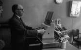 Stoppljus på systemet, 3 juni 1965

En man i kostym och glasögon står vid kassaapparaten bakom disken på Systembolaget tillsammans med en yngling. Den äldre mannen är i färd med att slå in något på kassaapparaten. På andra sidan disken syns ett vinställ.