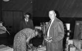 Drick vin gratis 31 juli 1965

Kvinna i klänning står framåtböjd och öppnar en trälåda med en hammare. Två män flankerar henne på ömse sidor. Den ene har en cigarett i mungipan och den andra håller en massa vinglas i vänster hand. Tavlor syns på golvet i bakgrunden.