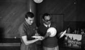 Drick vin gratis 31 juli 1965

Två män står med fat i händerna och betraktar dessa. Mannen till höger på bilden bär glasögon. I bakgrunden till höger syns tavlor som står på golvet samt ytterligare föremål.