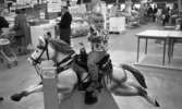 Stormarknaden, 31 juli 1965

Inne i Domus stormarknad. I förgrunden sitter en liten pojke på en automatisk leksakshäst med ett leksaksgevär i händerna. Åkturen kostar 50 öre vilket står bredvid på stolpen med myntintaget. I bakgrunden syns vuxna människor. Vissa skjuter kundvagnar framför sig. Varor till försäljning syns även som exempelvis cyklar, bord, tvättmaskin, frysbox etc.