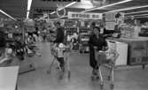 Stormarknaden, 31 juli 1965

Inne på Domus stormarknad. I förgrunden syns en kvinna och en man. Kvinnan går gången fram med fylld kundvagn och mannen står vid sin fyllda kundvagn. Gången kantas av hyllor med varor till försäljning såsom en cykel, leksaksbarnvagnar, vitvaror etc. En automatisk leksakshäst finns också i lokalen.