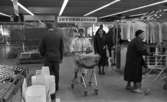 Stormarknaden, 31 juli 1965

I mitten i förgrunden inne på Domus stormarknad går en kvinna med en kundvagn. I vagnen sitter ett litet barn. Bredvid kvinnan står en man i kostym. En äldre dam står till höger med en kundvagn. I bakgrunden syns ytterligare två personer: två kvinnor och en man. I bakgrunden uppe vid taket hänger en skylt med texten 