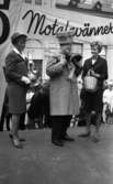 Knytkalaset 5 juli 1965.

Harald Aronsson stående på en scen med krona på huvudet och två kattungar i famnen. Den ena av de två värdinnorna på ömse sidor om honom håller i en prispropeller och en flagga. Den andra värdinnan håller i en korg. Publik i bakgrunden.