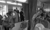 Damfrisörskor 8 juli 1965.

I förgrunden klipper en damfrisörska en ung pojke. I spegeln i bakgrunden reflekteras bilden. Där syns också andra människor passera salongen.