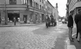 Hästar i stan, 8 juli 1965.

Man kör arbetshäst och vagn genom stan. Bredvid på trottoaren står en grupp unga män och samtalar.