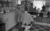 Damfrisörskor 8 juli 1965.

Frisörska klipper ung man. I spegeln syns en fotograf som tar bilder med en kamera.