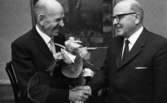 Trotjänare avtackad  7 december 1965

Två äldre herrar i mörka kostymer med vita skjortor och slipsar. Den ena herrn bär ett par svarta glasögon. Den ena herrn avtackar den andre med en bukett blommor. I bakgrunden hänger en stor tavla på väggen.