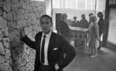 Club 700, 20 augusti 1965

I förgrunden står en man i mörk kostym, vit skjorta och slips. Han håller högra handen mot väggen. I bakgrunden står sju personer samlade runt en spelautomat. En entrédörr finns till höger om dem.