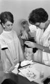 Frisörskan 2 september 1965

En damfrisörska (lärarinna) samt en hårfrisörskeelev står vid en frisördocka. Båda har vita arbetsrockar på sig. Lärarinnan kammar dockans hår och eleven tittar på.
På arbetsbordet ligger en broschyr med texten 