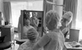 Frisörskan 2 september 1965

En äldre dam iförd skyddskläder sitter framför en spegel inne i en salong. En hårfrisörskeelev är i full färd med att kamma hennes hår. Bredvid till höger sitter en annan dam med håret i en torkhuv. Vid henne står ett skåp med borstar och hårvårdsredskap. Ett till skåp med lådor står längre till höger i bakgrunden. Till vänster skymtar två andra elever i vita arbetsrockar samt ett skåp med lådor och en stol.