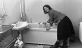 Hemma hos Karasko, 24 december 1965

En kvinna klädd i blus, kjol samt stövlar duschar badkaret i ett badrum. I rummet finns även handfat, tvättho samt ett element. En korg med en pappkasse i  står på golvet. En matta ligger även på golvet.