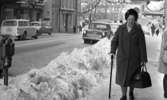 Halkiga pensionärer, 18 december 1965

En äldre dam- en pensionär- kommer gående på trottoaren en vinterdag på Drottninggatan. Hon är klädd i mörk lång kappa, pälsmössa med märke, mörka strumpor och skor på fötterna. I höger hand håller hon en käpp och i vänster hand en stor väska. Bredvid henne längs med trottoaren löper en stor, uppskottad snöhög. Längre fram vid trottoarkanten står två parkeringsautomater. Bilar åker på gatan i bakgrunden. Ytterligare personer promenerar i bakgrunden. Uppe i luften över gatan hänger juldekorationer.