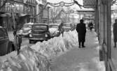 Halkiga pensionärer, 18 december 1965

I förgrunden syns en manlig pensionär med ryggen mot kameran. Han går på trottoaren på Drottninggatan. Vid sidan av trottoarkanten finns en snövall. Ur denna sticker parkeringsmätare upp. Bilar står parkerade längs med gatan invid trottoarkanten. Flera människor syns i bakgrunden. Längre fram på gatan hänger en skyltar med texterna 