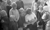 Inskrivning barngymnastik 15 september 1965

Barnen som ska skrivas in samlas runt lärarinnan, Inga Pettersson, tillsammans med några föräldrar. Lärarinnan, Inga Pettersson, sitter på en bänk och antecknar och har på bilden vänt sig bakåt för att titta på ett av barnen.
