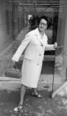 Modebilder 23 april 1966

En kvinna iförd modekläder: en vit kappa, ljusa lågklackade skor och med en ljus handväska i höger hand är på väg ut genom en glasdörr. Hon håller i dörren med vänster hand. Hon bär glasögon samt har ett pärlhalsband om halsen.