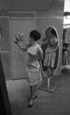 Modebilder 23 april 1966

En mannekäng intar en pose klädd i ljus figursydd kort klänning och svarta pumps håller en slags bukett i sin högra hand och tittar på den. Hon står framför en spegel. Reflexionen av hennes baksida syns i spegeln. I bakgrunden syns ställ med blusar som hänger på galgar.
