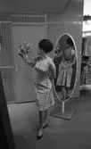 Modebilder 23 april 1966

En mannekäng intar en pose klädd i ljus figursydd kort klänning och svarta pumps håller en slags bukett i sin högra hand och tittar på den. Hon står framför en spegel. Reflexionen av hennes baksida syns i spegeln. I bakgrunden syns ställ med blusar som hänger på galgar.