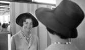 Modebilder 23 april 1966

En leende mannekäng klädd i mörk, vidbrättad hatt, ljus kavaj och svartvitt halsband tittar in i en spegel. Reflexionen från spegeln visar hennes ansikte och lite av överkroppen.