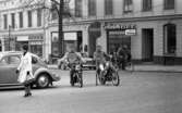 Mopedister 5 maj 1966

Två unga pojkar åker på mopeder på en gata i centrala Örebro. De passerar förbi en vägskylt. Med ryggen vänd åt dem går en kvinna på gatan. Hon är klädd i en vit kappa, svart halsduk, svarta skor, svarta handskar och med en svart handväska i handen. Hon bär glasögon. En äldre herre cyklar i bakgrunden. Bilar syns på gatan.