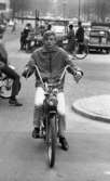 Mopedister 5 maj 1966

En ung pojke sitter på en moped på en gata i centrala Örebro. Han är klädd i jacka, byxor och skor. Lite i bakgrunden till vänster sitter hans kompis också på en moped. Längre i bakgrunden står barn och vuxna samlade kring ett stånd. I närheten står bilar parkerade.
